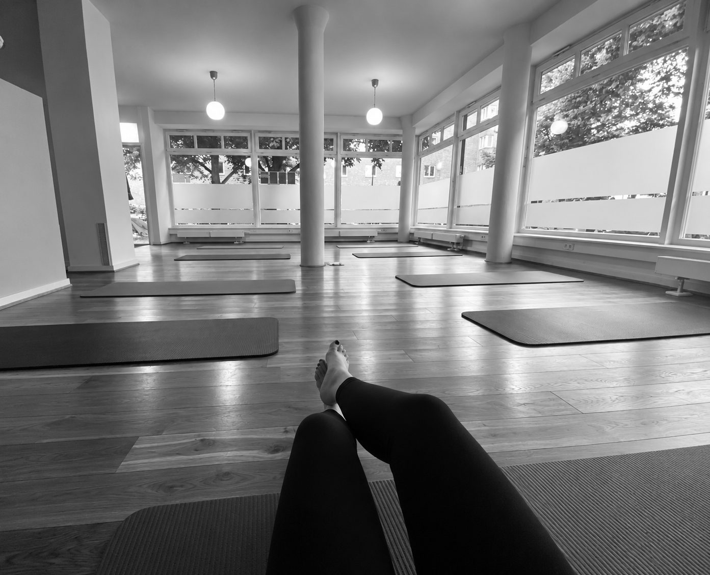 Blick über die eigenen ausgestreckten Beine auf eine Trainingsmatte in einem hellen Pilates-Studio mit Holzboden, weiteren Matten und großen Fenstern.